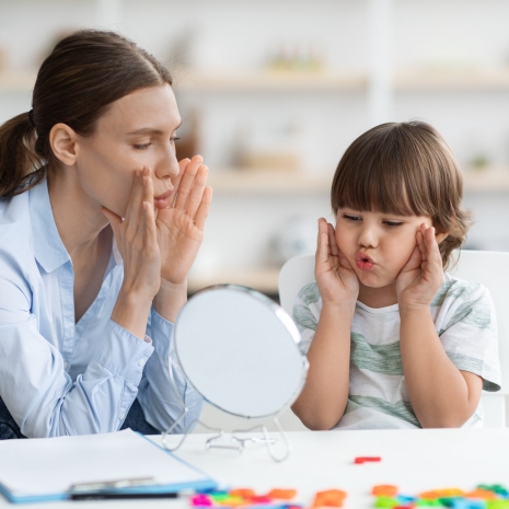 Cute little boy with speech problems practicing with therapist, doing practice and looking at mirror, free space